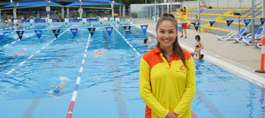 Female lifeguard with long brown hair wearing bright yellow and red long sleeve top standing in front of a blue outdoor pool