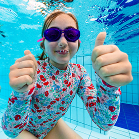 Kid swimming under water with floral pink bathing suit holding two thumbs up to camera and smiling.