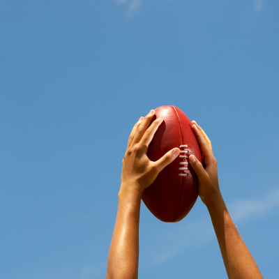 Two hands reaching up to catch an AFL football with blue sky behind