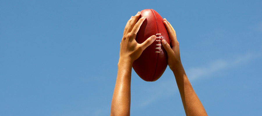 Two hands reaching up to catch an AFL football with blue sky behind