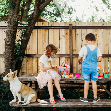 A young boy and girl standing on a wooden table in their backyard playing with easter eggs and toys with their dog next to them