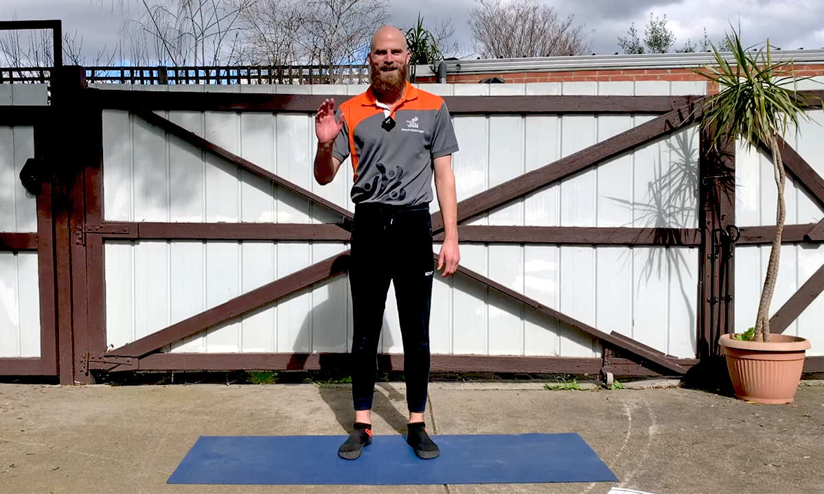 Gym Instructor Leigh standing in front of a fence at home and standing on a yoga mat waving and smiling